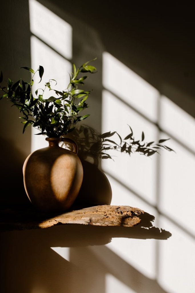 A minimalist vase containing greenery placed on a wooden surface with dramatic sunlight and shadows indoors.